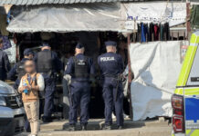 Grupa policjantów w mundurach rozmawia z osobami w stoisku na targowisku, w pobliżu zaparkowanego samochodu.