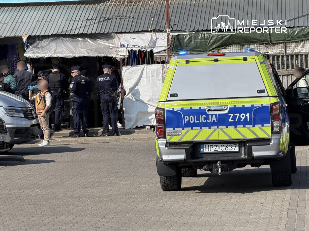 Grupa policjantów prowadzi interwencję przed straganem na targowisku, a w tle widać zaparkowany radiowóz.
