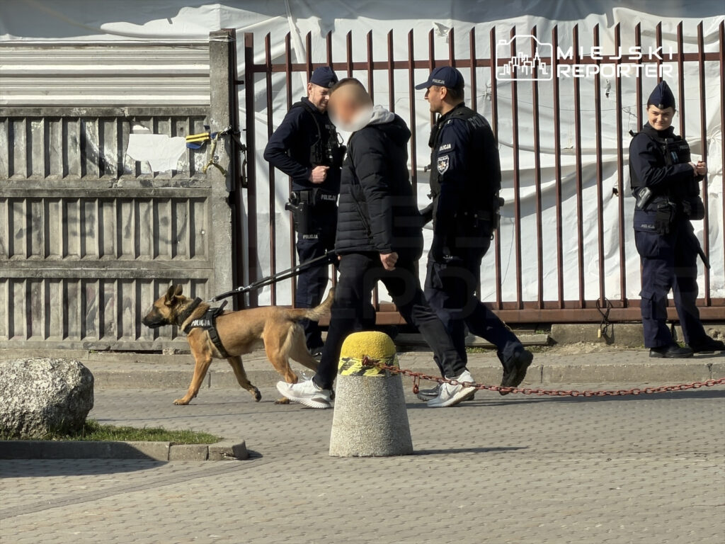 Policjant prowadzi psa na smyczy, idąc obok grupy funkcjonariuszy w miejskim otoczeniu.