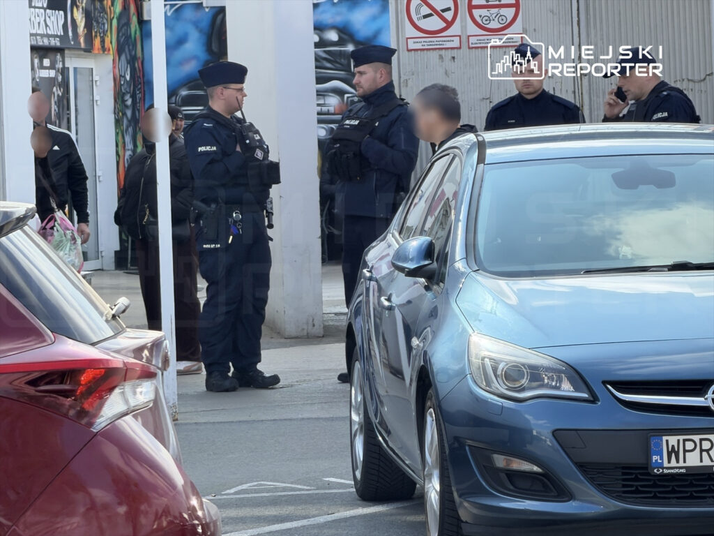 Grupa policjantów w mundurach rozmawia z osobami stojącymi w pobliżu graffiti na ścianie, w tle zaparkowane samochody.