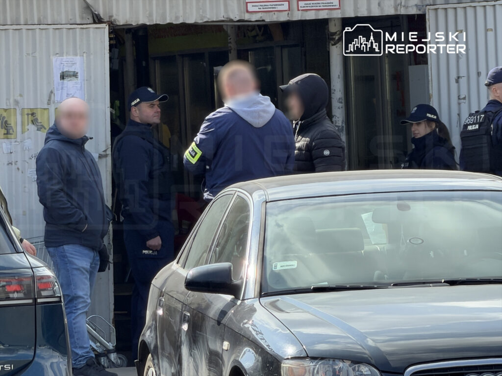 Grupa mężczyzn rozmawia przed budynkiem, w którym znajdują się drzwi i plakaty informacyjne, w otoczeniu zaparkowanych samochodów.