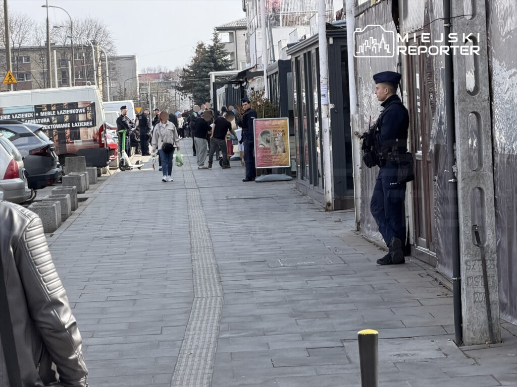 Grupa osób z zakupami spaceruje po chodniku, obok stoi policjant w mundurze, a w tle widoczne są pojazdy.