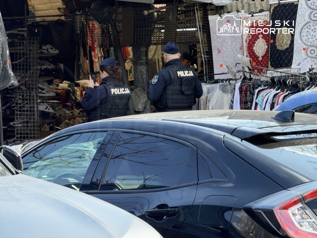 Dwóch policjantów w mundurach rozmawia w zatłoczonym targowisku, w tle wystawy odzieżowe i obuwie.
