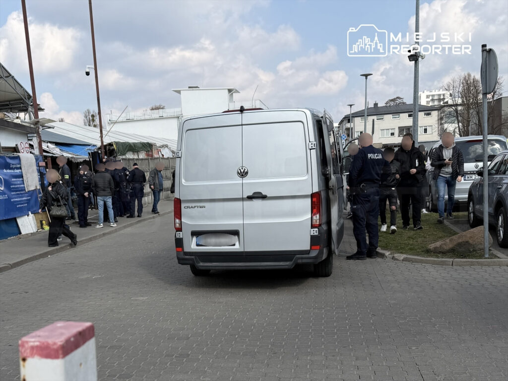 Grupa policjantów prowadzi interwencję w pobliżu straganów na targowisku, obok zaparkowanych samochodów.