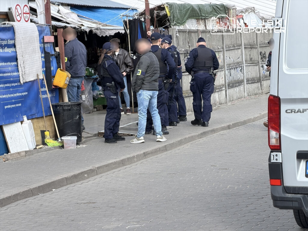 Grupa policjantów rozmawia z mężczyzną na chodniku przed straganem z towarami.