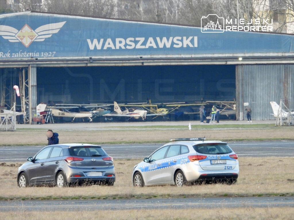Samoloty w hangarze na lotnisku, obok policyjny radiowóz i osoby pracujące na zewnątrz.