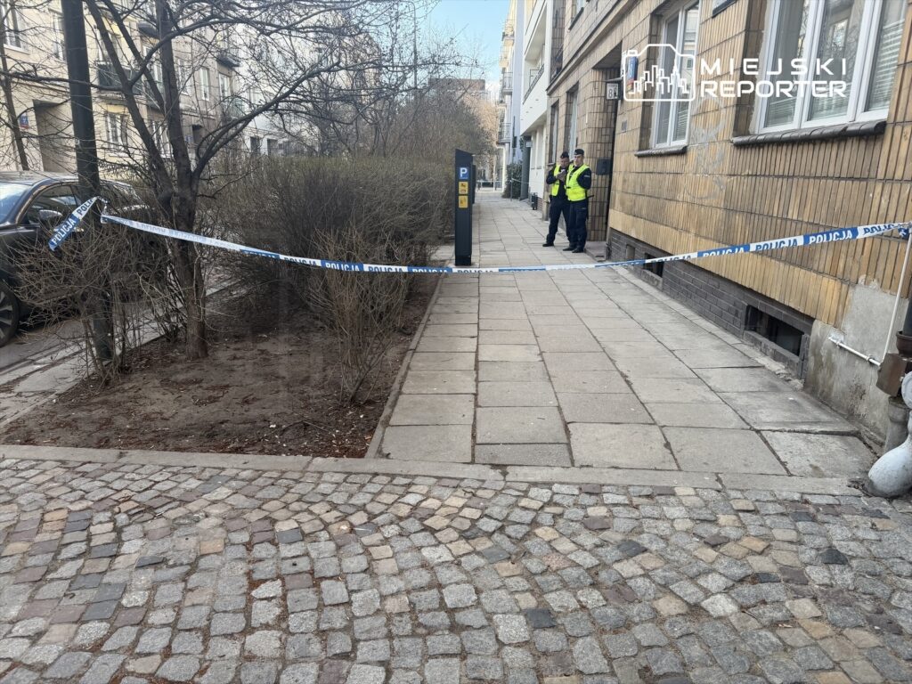 Policjanci zabezpieczają miejsce zdarzenia, przy oznakowanym taśmą obszarze na chodniku.