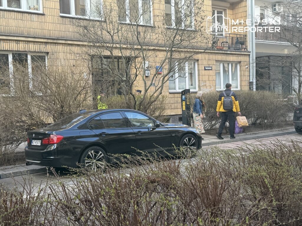 Mężczyzna w żółtej kurtce niesie torbę, idąc obok kobiety w długiej sukience wzdłuż ulicy, obok zaparkowanego czarnego samochodu.