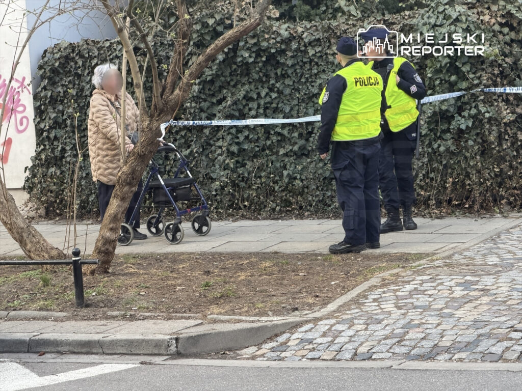 Starsza kobieta z chodzikiem stoi na chodniku obok policjantów w kamizelkach odblaskowych, rozmawiających przy ogrodzeniu.