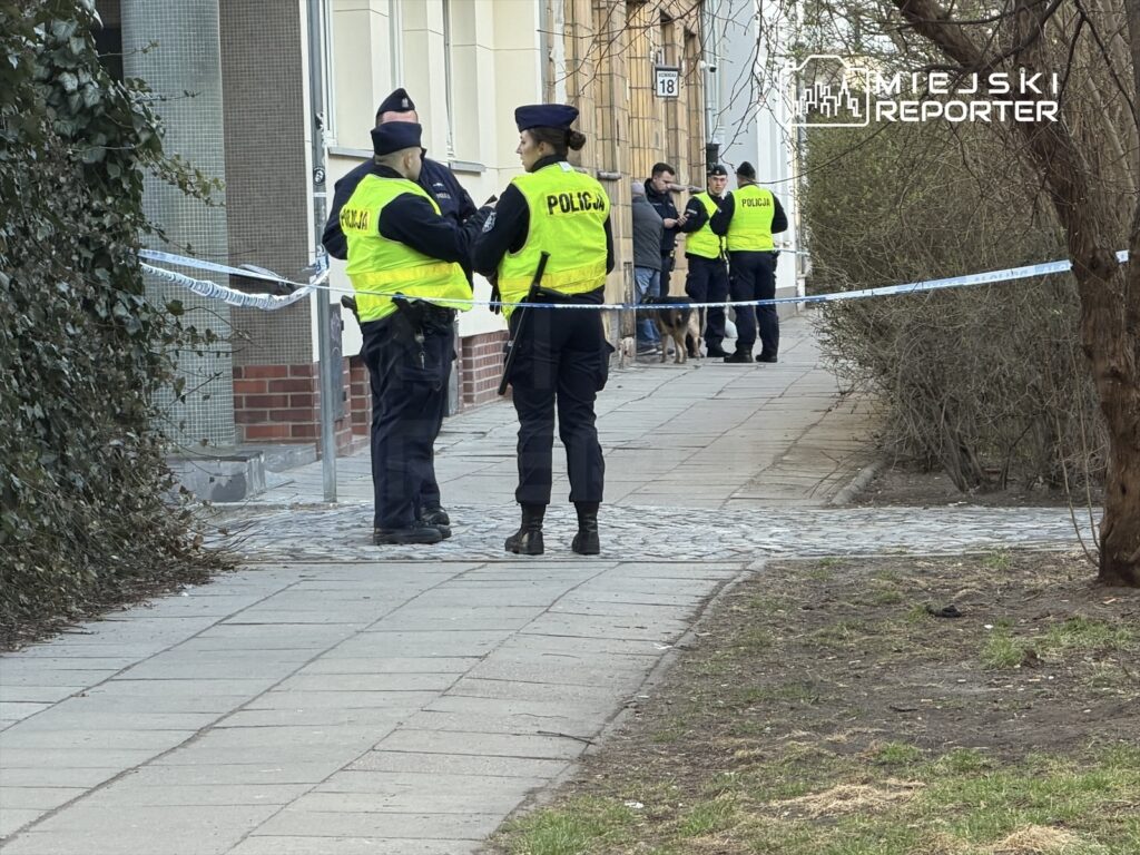 Grupa policjantów w kamizelkach odblaskowych rozmawia na chodniku, zabezpieczając miejsce zdarzenia taśmą.
