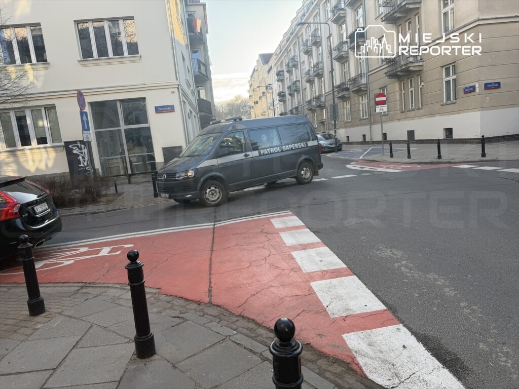 Samochód dostawczy oznaczony napisem "PATROL SAPERSKI" skręca w lewo na skrzyżowaniu, obok nowoczesnych budynków.