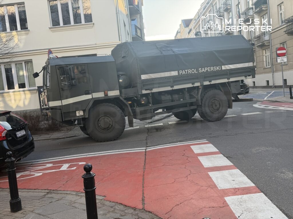 Ciężarówka wojskowa z napisem "PATROL SAPERSKI" skręca na skrzyżowaniu w mieście, obok zaparkowanego samochodu osobowego.