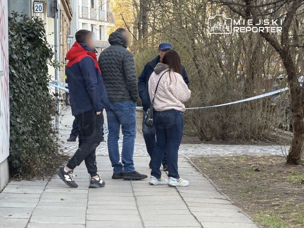 Grupa czterech osób rozmawia na chodniku, obok niebieskiej taśmy zabezpieczającej, w otoczeniu drzew i budynków.