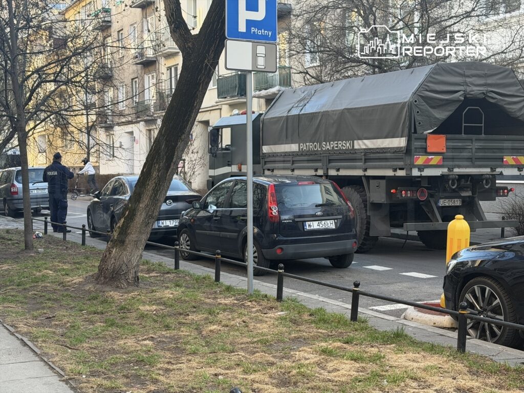 Policjant w mundurze obserwujący ruch na ulicy, obok przejeżdżający pojazd wojskowy z napisem "Patrol Saperski".