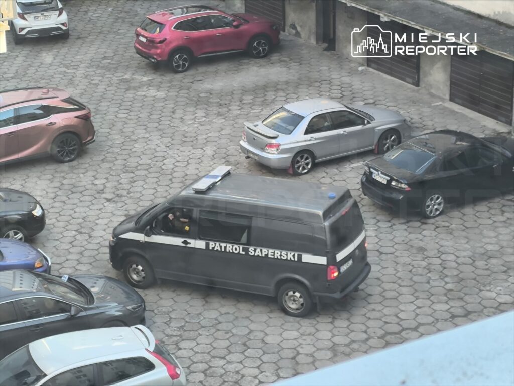 Patrol saperski porusza się po parkingu, w otoczeniu zaparkowanych samochodów różnych marek.