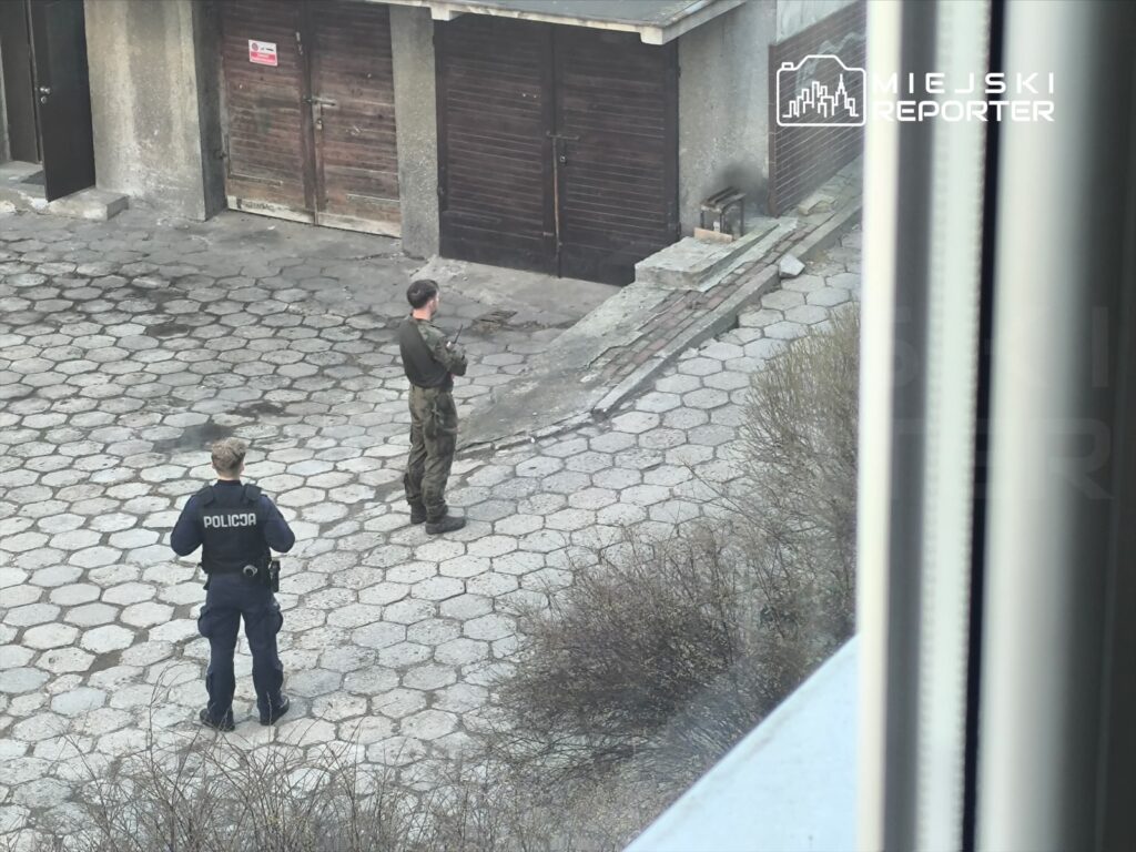 Policjant w mundurze stoi na placu, obok niego żołnierz z bronią, w tle zamknięte garaże.