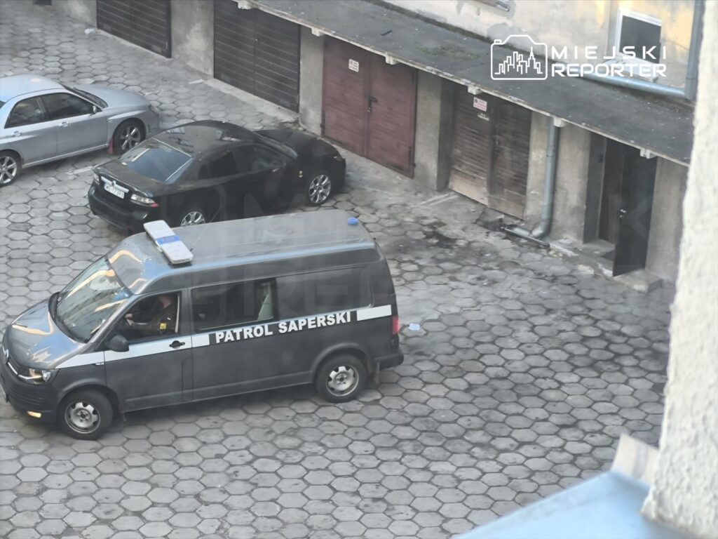 Patrol saperski zaparkowany na podwórku, obok dwóch samochodów osobowych, w otoczeniu garaży.