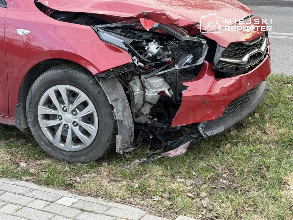 Czerwony samochód osobowy z uszkodzoną przednią częścią stoi na trawie obok chodnika.