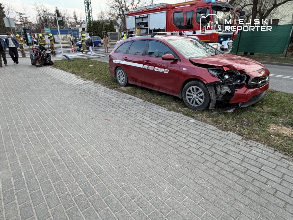 Czerwony samochód osobowy z uszkodzoną przodem stoi na poboczu, obok grupy strażaków i przechodniów.