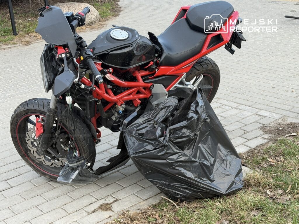 Motocykl z czerwoną ramą zaparkowany obok czarnej torby na śmieci na chodniku w miejskim otoczeniu.