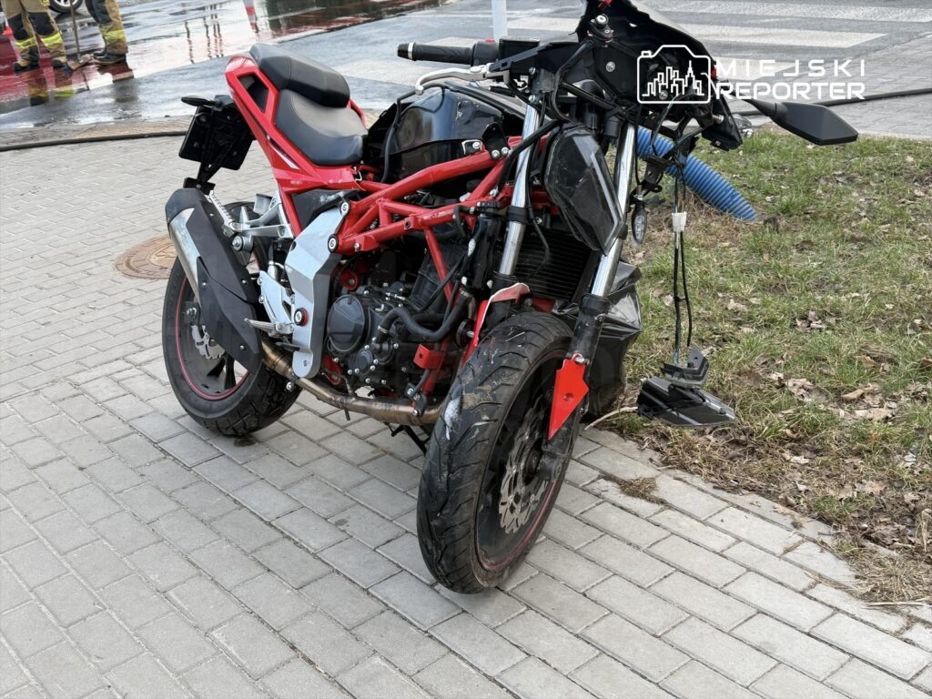 Motocykl o sportowym wyglądzie z czerwoną ramą stoi na chodniku, obok widać fragment trawnika i hydrant.