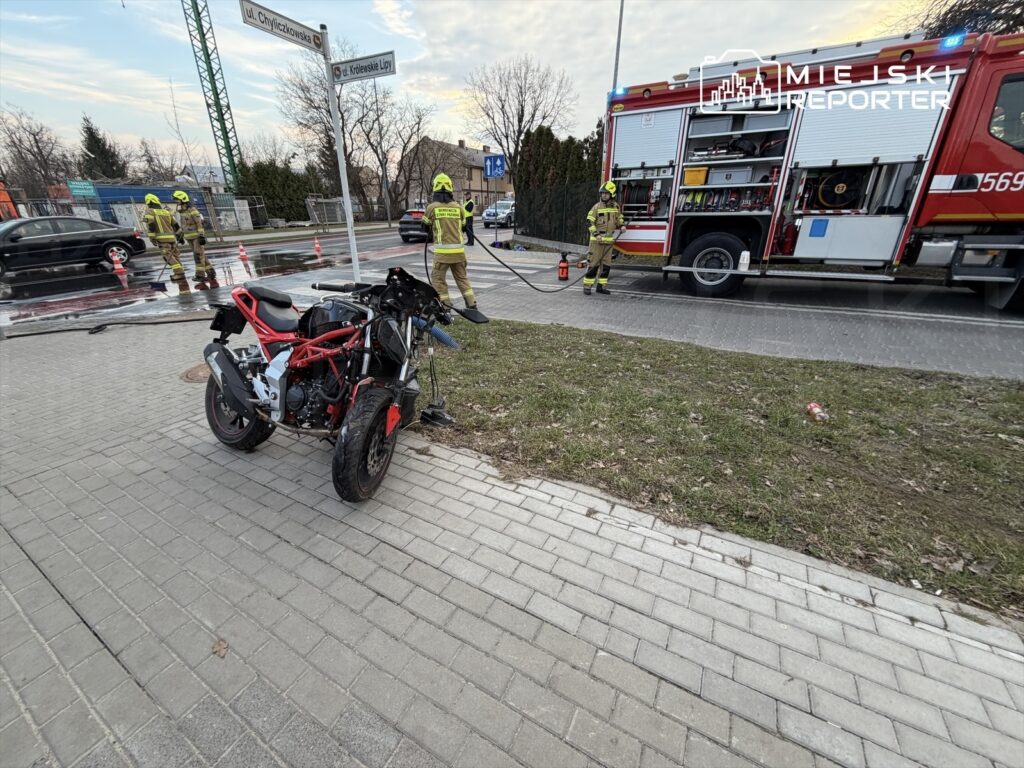 Motocykl z uszkodzoną przednią częścią stoi na chodniku, obok strażacy gaszący pożar w samochodzie.