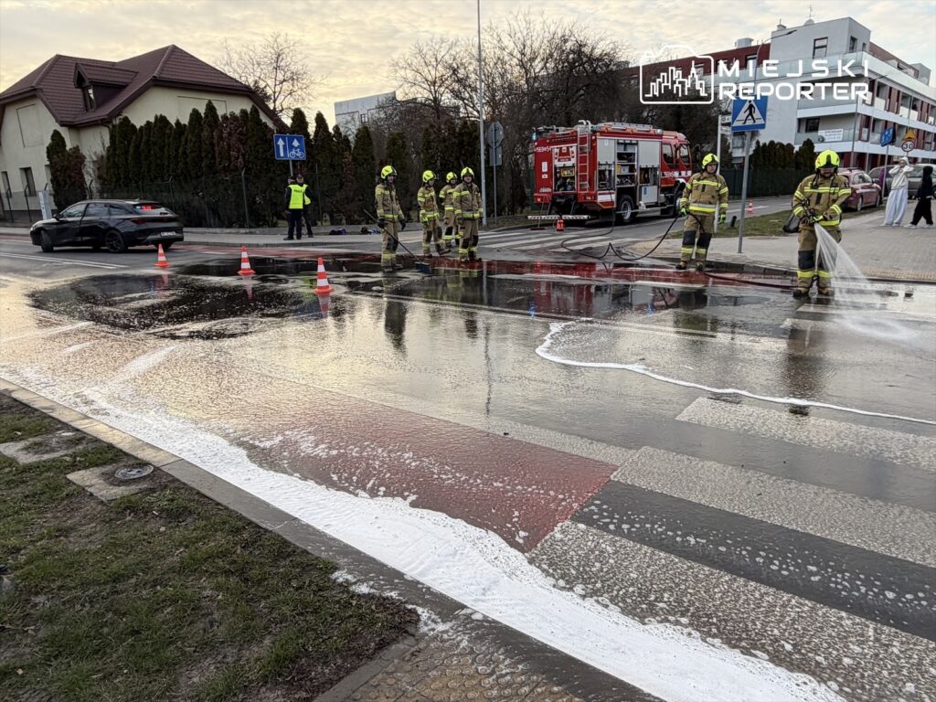 Grupa strażaków w mundurach używa węży strażackich do usuwania piany z jezdni na skrzyżowaniu.