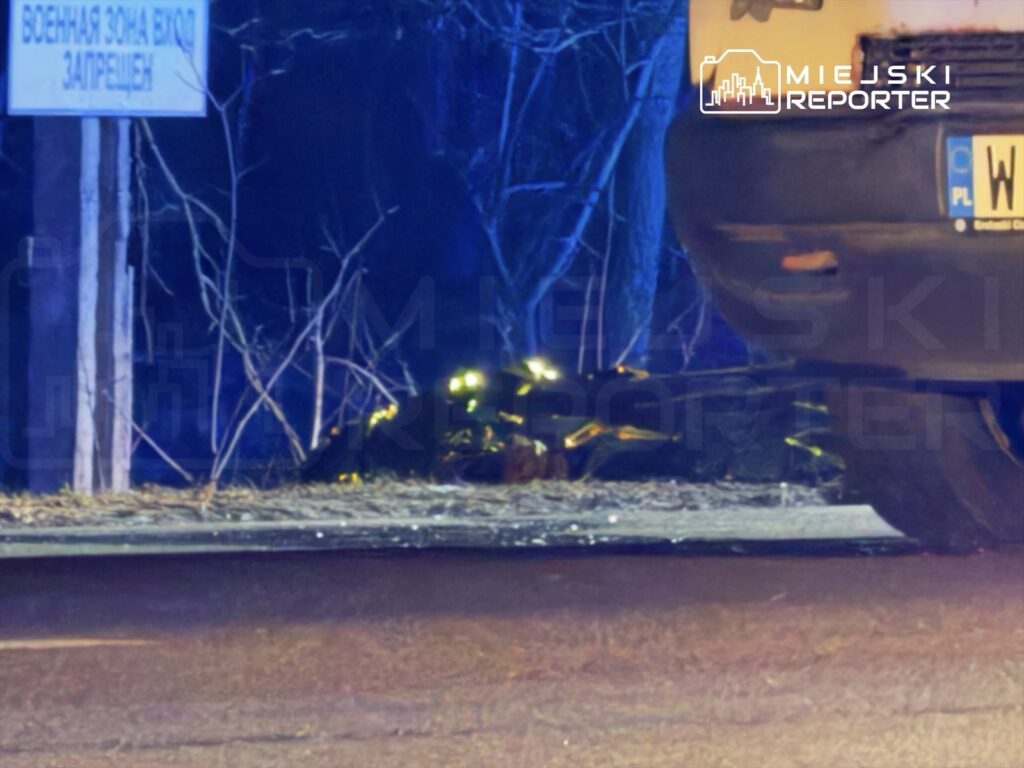 Na drodze widoczny jest nieoznakowany samochód z włączonymi światłami, obok leży uszkodzony pojazd w ciemnym otoczeniu.