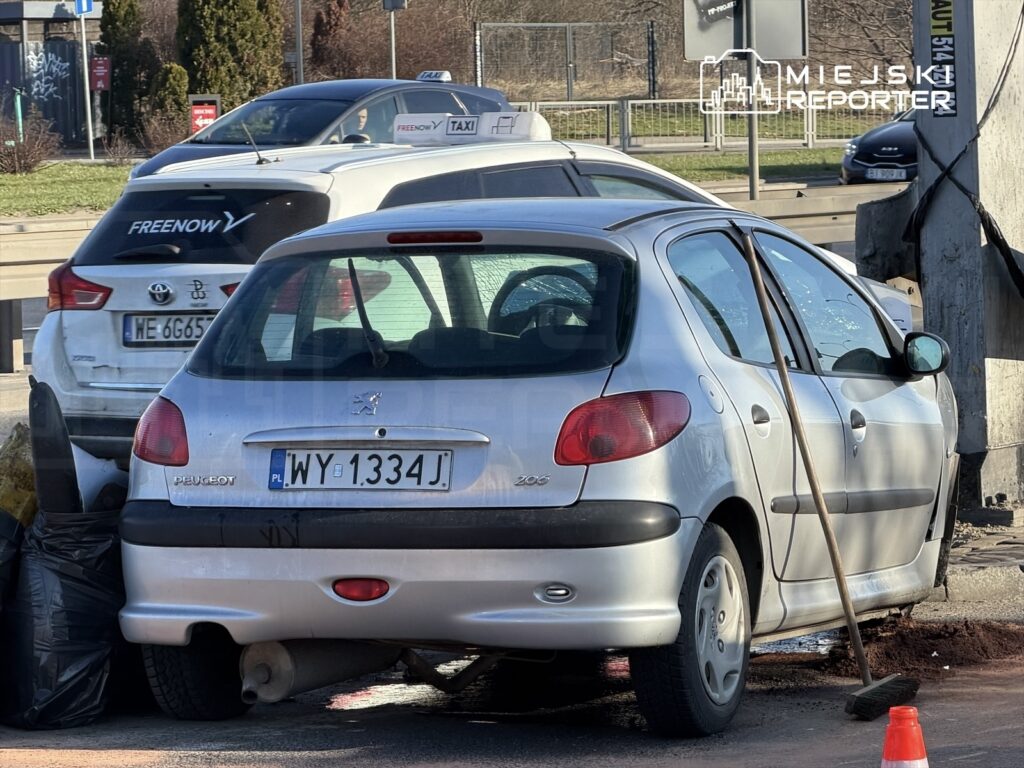 Srebrny Peugeot 206 zaparkowany w pobliżu stacji benzynowej, obok worka na śmieci i miotły.