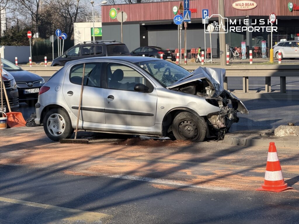 Uszkodzony samochód osobowy stoi na drodze, obok widoczne narzędzia do sprzątania i pachołki drogowe.