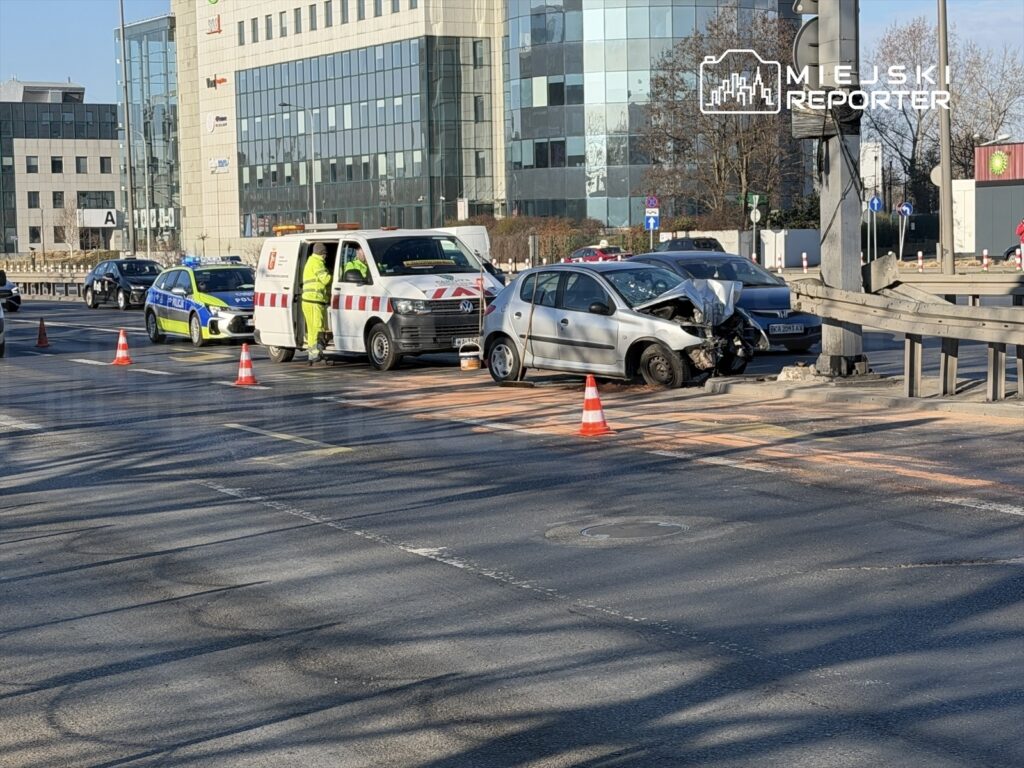 Na skrzyżowaniu ulic stoi uszkodzony samochód osobowy, obok niego zespół ratunkowy oraz radiowóz policyjny.