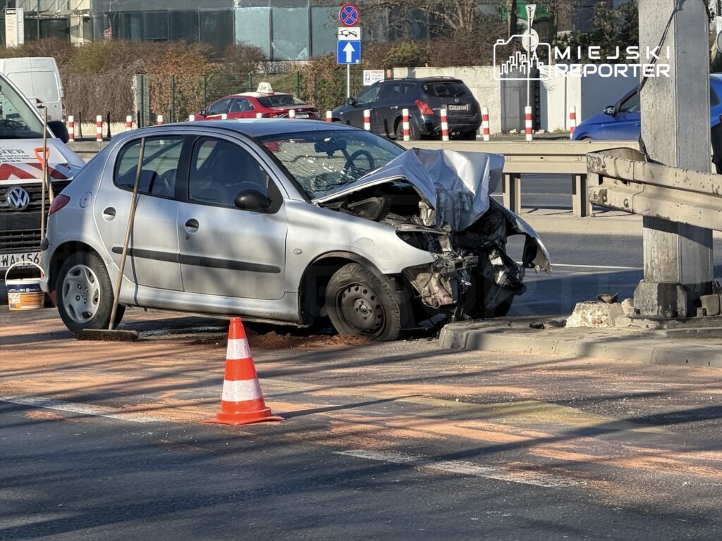 Uszkodzony samochód osobowy z wgniecioną maską stoi na jezdni obok bariery ochronnej, otoczony pachołkami drogowymi.