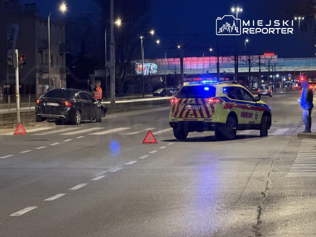 Czarny samochód osobowy stoi na poboczu drogi, obok policjant w kamizelce odblaskowej rozmawia z kierowcą, a w tle widać ambulans.