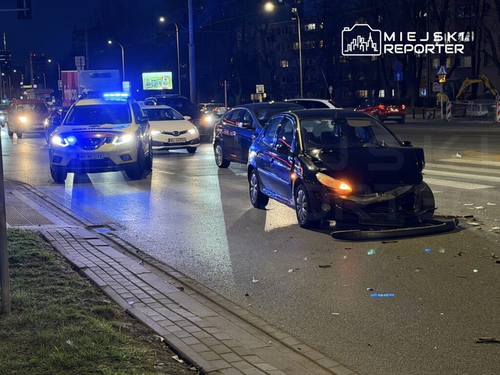 Czarny samochód osobowy z uszkodzoną przednią częścią stoi na jezdni, obok niego policyjny radiowóz z włączonymi sygnałami świetlnymi.