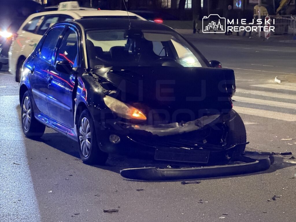 Czarny samochód osobowy z uszkodzoną przednią częścią stoi na drodze, obok widać inne pojazdy w ruchu.
