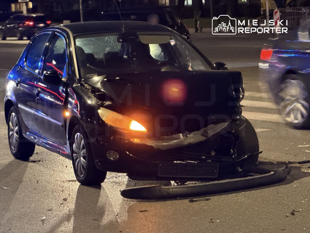 Czarny samochód osobowy z uszkodzoną przednią częścią stoi na ulicy, obok widoczne reflektory innych pojazdów.