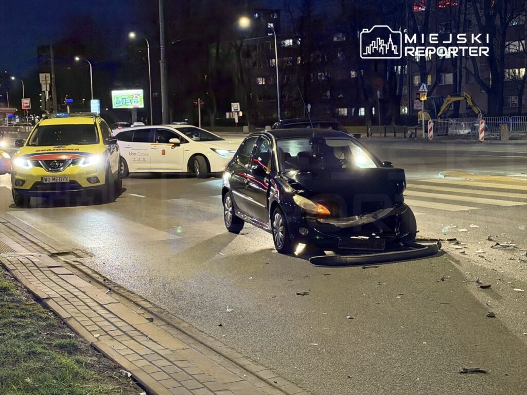 Na ruchliwej ulicy nocą, uszkodzony czarny samochód stoi na pasie drogowym, obok niego zaparkowany żółty pojazd ratunkowy.