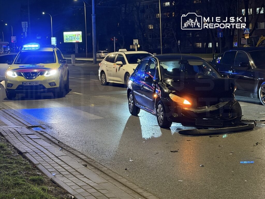 Czarny samochód osobowy z uszkodzoną przednią częścią stoi na drodze, obok policyjny radiowóz z włączonymi sygnałami świetlnymi.