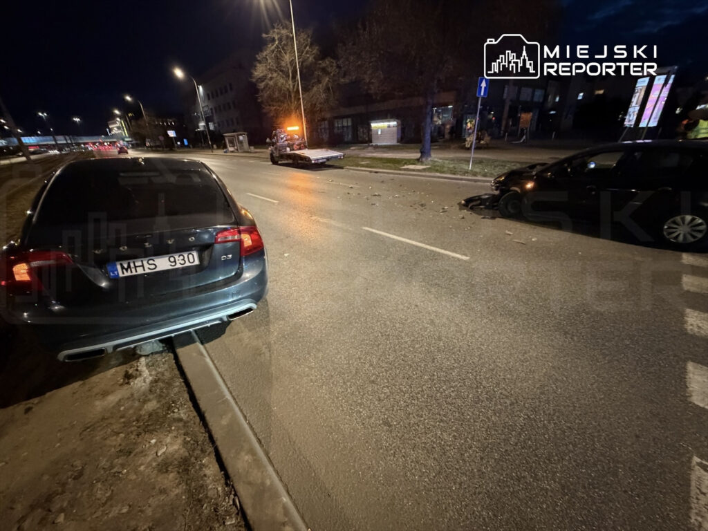 Czarny samochód osobowy z uszkodzoną przednią częścią stoi na poboczu, obok nieoznakowany wóz pomocy drogowej w nocy.