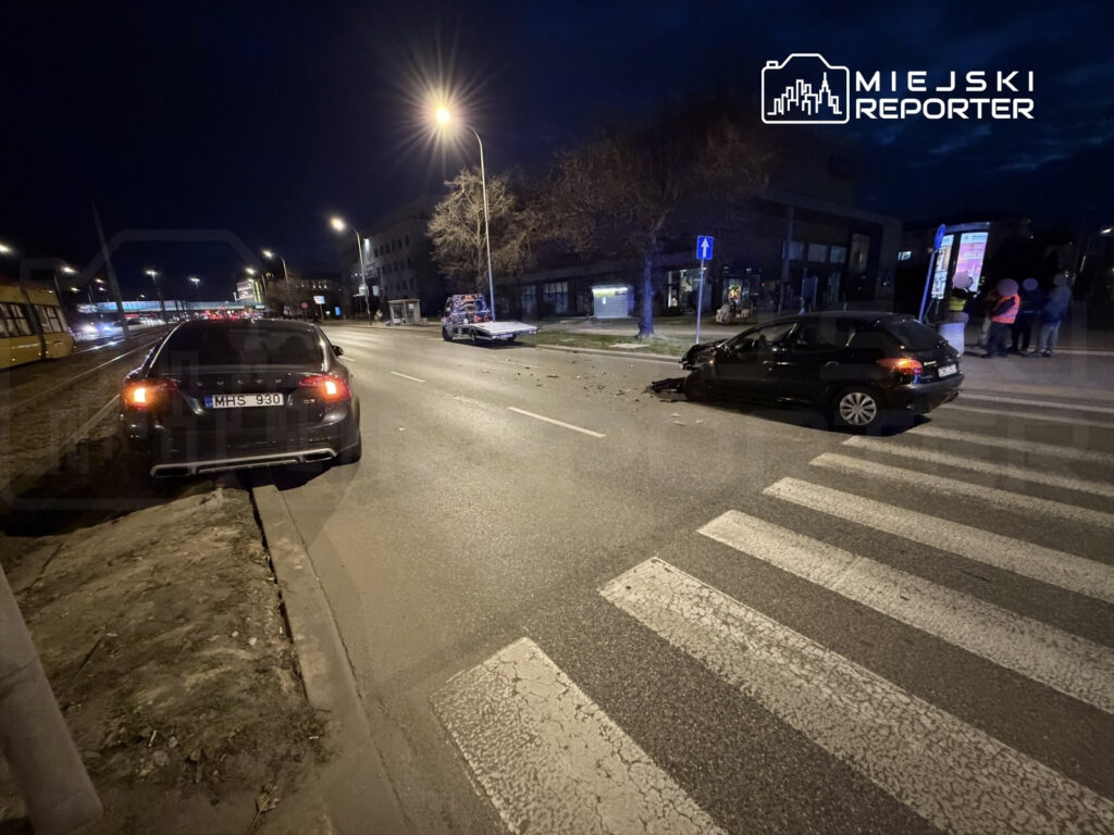 Dwa uszkodzone samochody po kolizji na ulicy nocą, obok przystanku autobusowego, z przechodniami w tle.