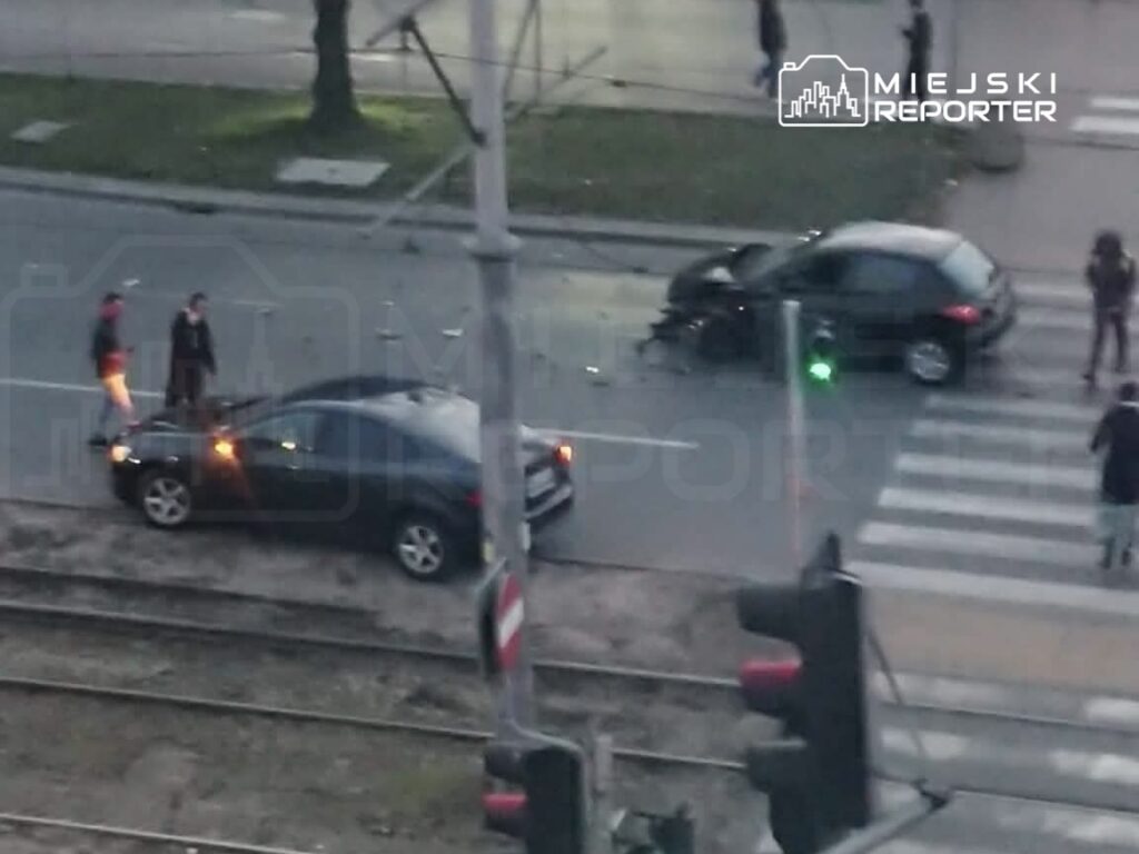 Dwie osoby rozmawiają obok uszkodzonego samochodu na skrzyżowaniu, w tle widać sygnalizację świetlną.