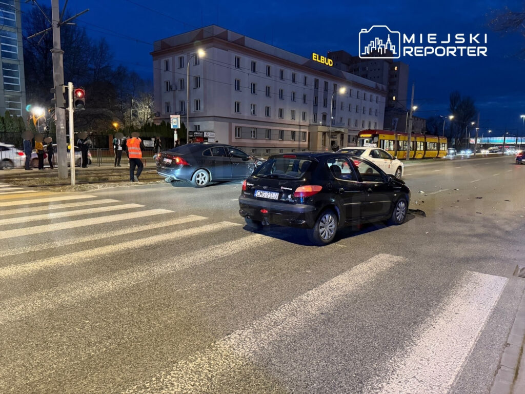Czarny samochód osobowy stoi na pasach, obok widać grupę ludzi oraz pracownika w pomarańczowej kamizelce.