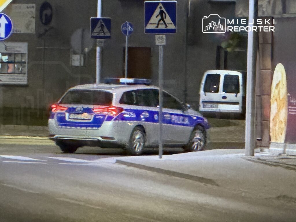Policyjny radiowóz zatrzymany na oświetlonej ulicy, obok znaki drogowe i zaparkowany biały furgon.