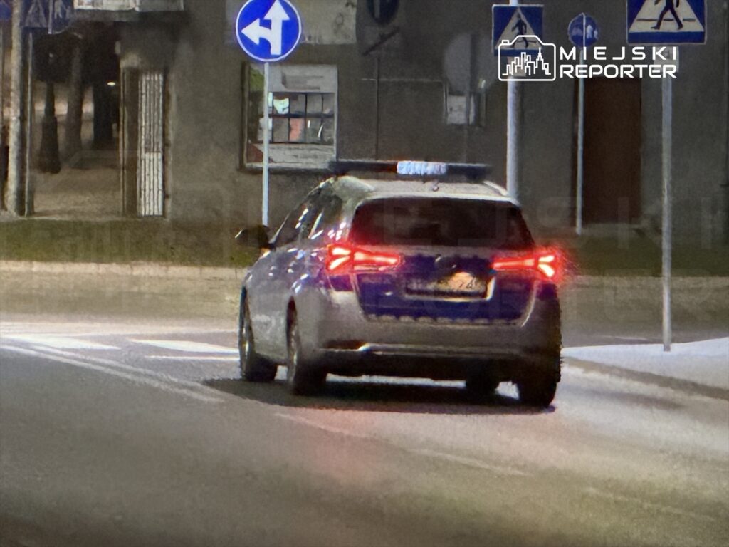 Radiowóz policyjny skręca w lewo na skrzyżowaniu w mieście, oświetlony przez latarnie uliczne.