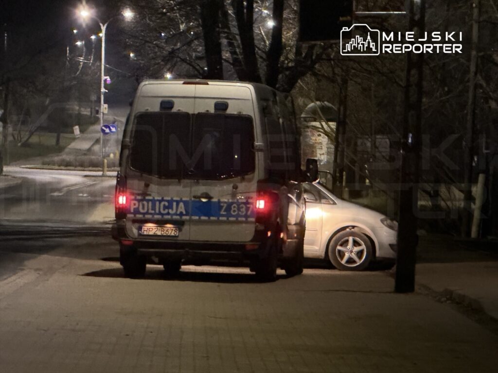 Policyjny radiowóz zaparkowany na ulicy nocą obok zaparkowanego srebrnego samochodu.