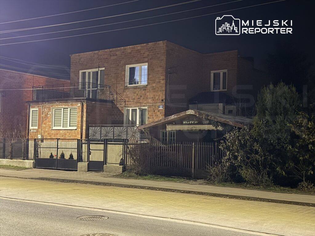 Czerwony budynek z cegły z balkonem i przeszklonymi oknami, otoczony ogrodzeniem, nocą przy ulicy.