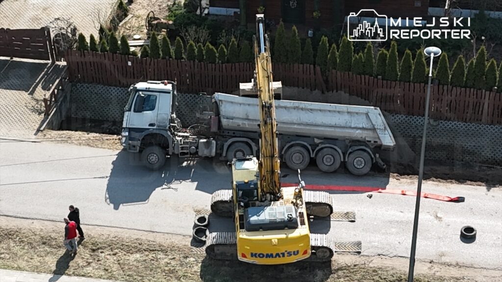 Dwa mężczyźni spacerują obok ciężkiego sprzętu budowlanego na drodze, w tle widoczny jest dom i ogrodzenie.