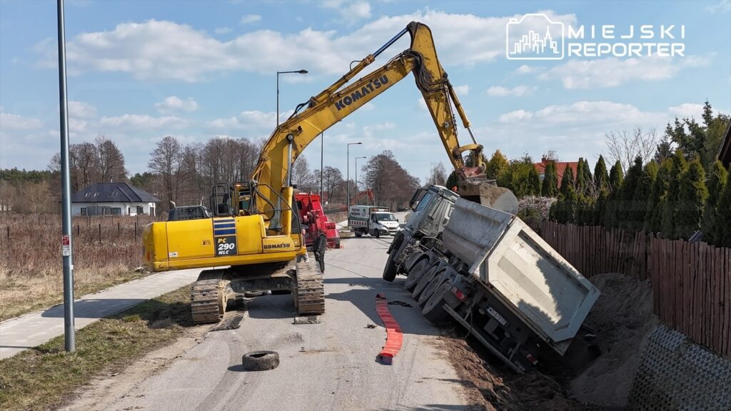 Koparka podnosi przód przewróconego ciężarowego pojazdu na drodze, obok widoczny jest samochód pomocniczy.