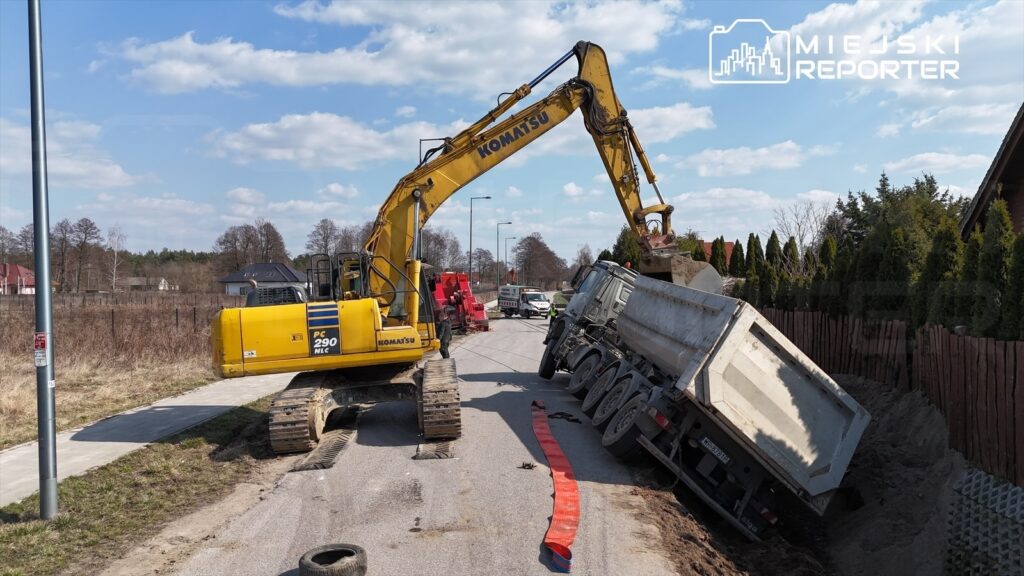 Żółty koparka podnosi przewróconą ciężarówkę na poboczu drogi, obok widoczne są drzewa i ogrodzenia.