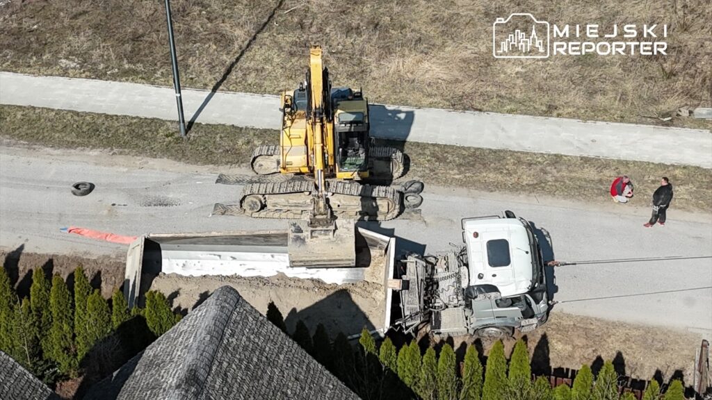 Koparka podnosi ziemię obok załadowanego ciężarowego pojazdu na drodze, a dwie osoby obserwują prace.